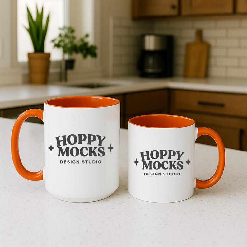May include: Two white ceramic mugs with orange interiors and handles. The mugs have the text "HOPPY MOCKS DESIGN STUDIO" printed in black. One mug is larger than the other. The mugs are on a white countertop.