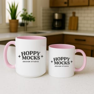 May include: Two white ceramic mugs with pink interiors and handles. The mugs have the text "HOPPY MOCKS DESIGN STUDIO" in black. One mug is larger than the other. The mugs are on a white countertop.
