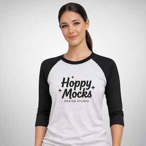 Op de afbeelding: Een baseball-stijl t-shirt met een witte body en zwarte mouwen. Het shirt heeft de tekst "Hoppy + Mocks DESIGN STUDIO" in een zwart, gestileerd lettertype. Het model draagt het shirt.