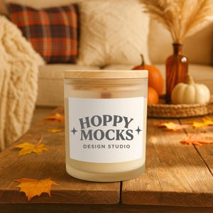 Peut inclure: Une bougie en verre dépoli avec un couvercle en bois sur une table en bois rustique. L'étiquette blanche de la bougie indique "HOPPY MOCKS DESIGN STUDIO". Des feuilles d'automne et des citrouilles décorent la table, créant une ambiance automnale.