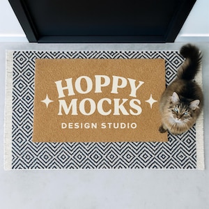 Könnte beinhalten: Eine rechteckige Fußmatte mit dem Text "HOPPY MOCKS DESIGN STUDIO" in einer Retro-Schriftart. Die Matte ist beige mit einem schwarz-weißen geometrischen Muster. Eine flauschige Katze sitzt rechts von der Matte.