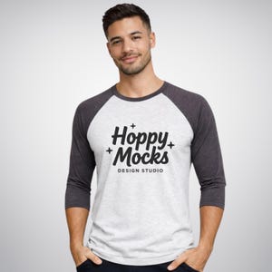 Op de afbeelding: Een raglan baseball t-shirt met een witte body en donkergrijze mouwen. Het shirt heeft de tekst "Hoppy Mocks DESIGN STUDIO" in een zwart, gestileerd lettertype. Het model draagt het shirt.
