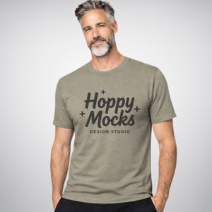 Può includere: T-shirt verde oliva con la scritta "Hoppy Mocks DESIGN STUDIO" in nero. La maglietta a maniche corte ha lo scollo tondo. Il modello indossa pantaloni neri.