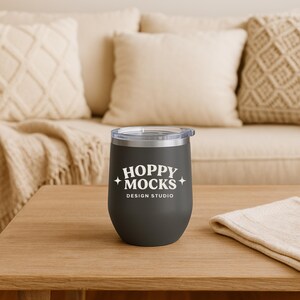 May include: A dark gray stemless tumbler with a silver rim and a clear lid. The tumbler has the text "HOPPY MOCKS DESIGN STUDIO" in white. The tumbler is on a wooden table with a beige cloth.