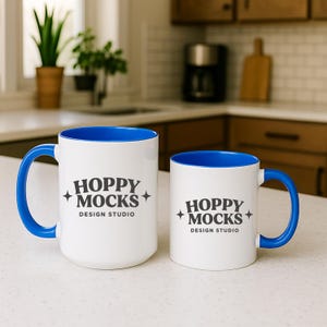 May include: Two white ceramic mugs with blue interiors and handles. The mugs have the text "HOPPY MOCKS DESIGN STUDIO" printed in gray. One mug is larger than the other. The mugs are on a white countertop.