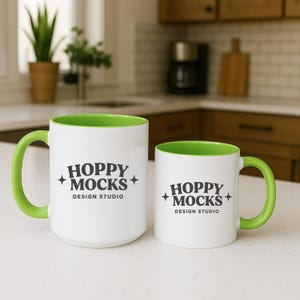 May include: Two white ceramic mugs with green interiors and handles. The mugs have the text "HOPPY MOCKS DESIGN STUDIO" printed in black. One mug is larger than the other.