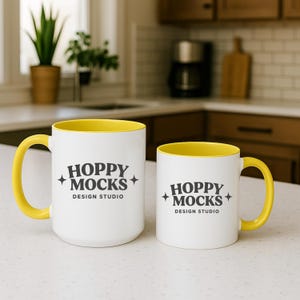 May include: Two white ceramic mugs with yellow interiors and handles. The mugs have the text "HOPPY MOCKS DESIGN STUDIO" printed in black. One mug is larger than the other, and both are on a white surface.