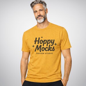 May include: Mustard yellow t-shirt featuring the text "Hoppy + Mocks DESIGN STUDIO" in black script. The short-sleeved shirt has a crew neck. The model is wearing black trousers. The background is a plain light grey.