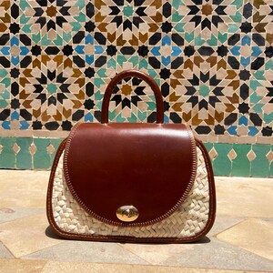 May include: A brown leather and woven straw handbag with a top handle and a gold-colored clasp. The bag has a rounded shape and a flap closure. The background features a colorful mosaic tile wall.