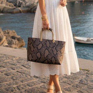 May include: A white eyelet midi dress with short sleeves and a V-neck. The dress is paired with a brown snakeskin tote bag with gold handles and beige sandals. The background features a coastal town.