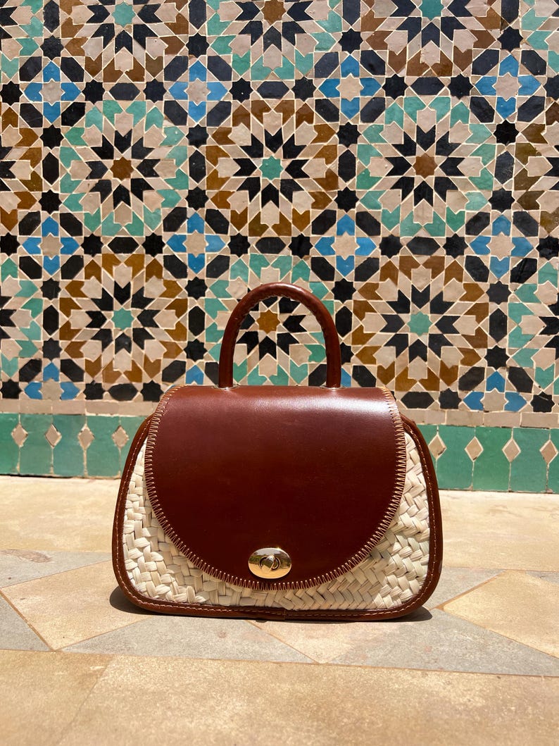 May include: A brown leather and woven straw handbag with a top handle and a gold-colored clasp. The bag has a rounded shape and a flap closure. The background features a colorful mosaic tile wall.
