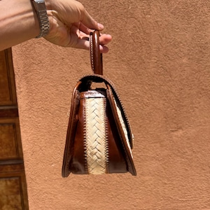 May include: A brown leather handbag with a woven cream-colored center panel. The bag has a top handle and a rectangular shape with a flap closure. The bag is held by a person.
