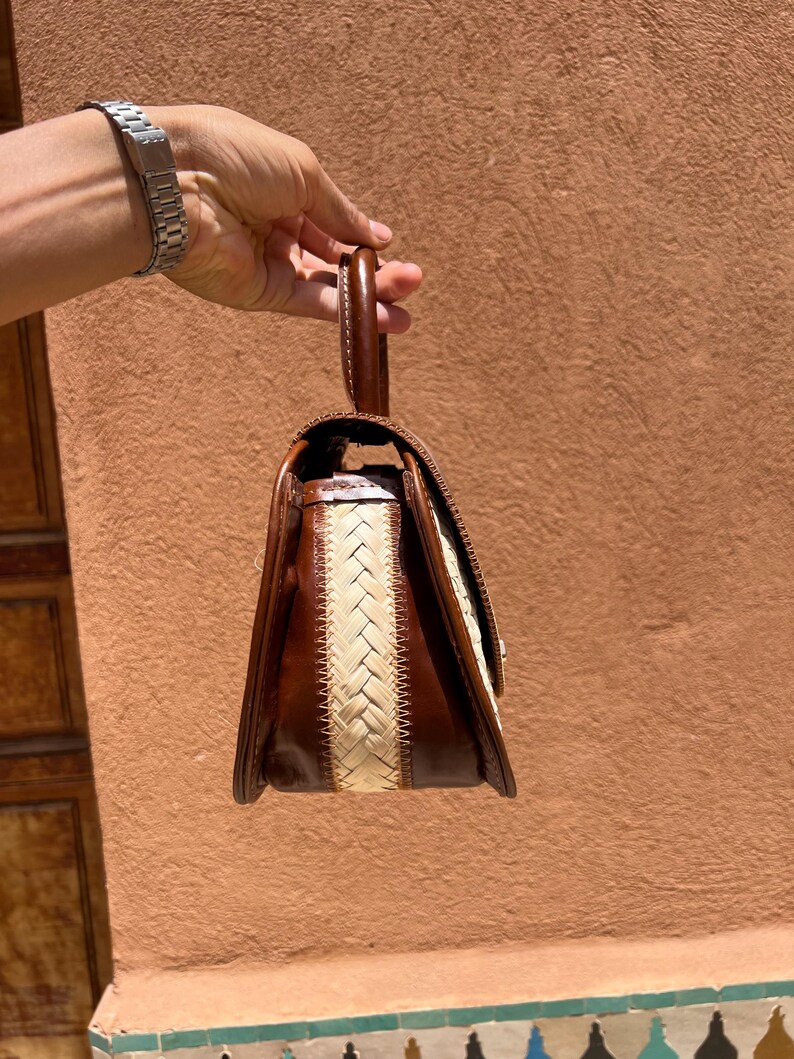 May include: A brown leather handbag with a woven cream-colored center panel. The bag has a top handle and a rectangular shape with a flap closure. The bag is held by a person.