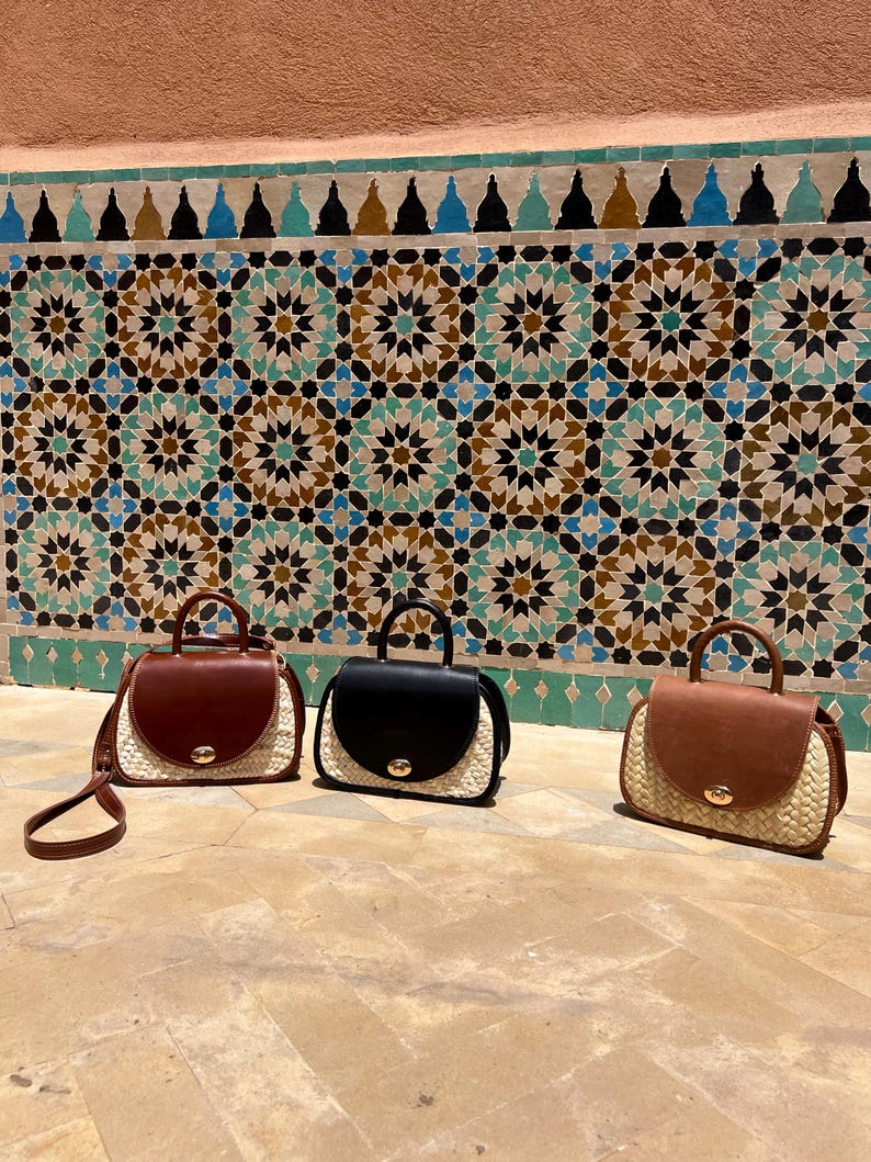 May include: Three handbags with a top handle and a flap closure. The bags have a woven straw base and a leather top in brown and black. The bags are displayed in front of a decorative tile wall.