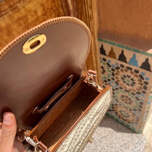 May include: Open view of a woven straw crossbody bag with brown leather trim and a brown leather interior. The bag has a gold-colored clasp and a long, adjustable strap. The bag is partially open, revealing the interior.