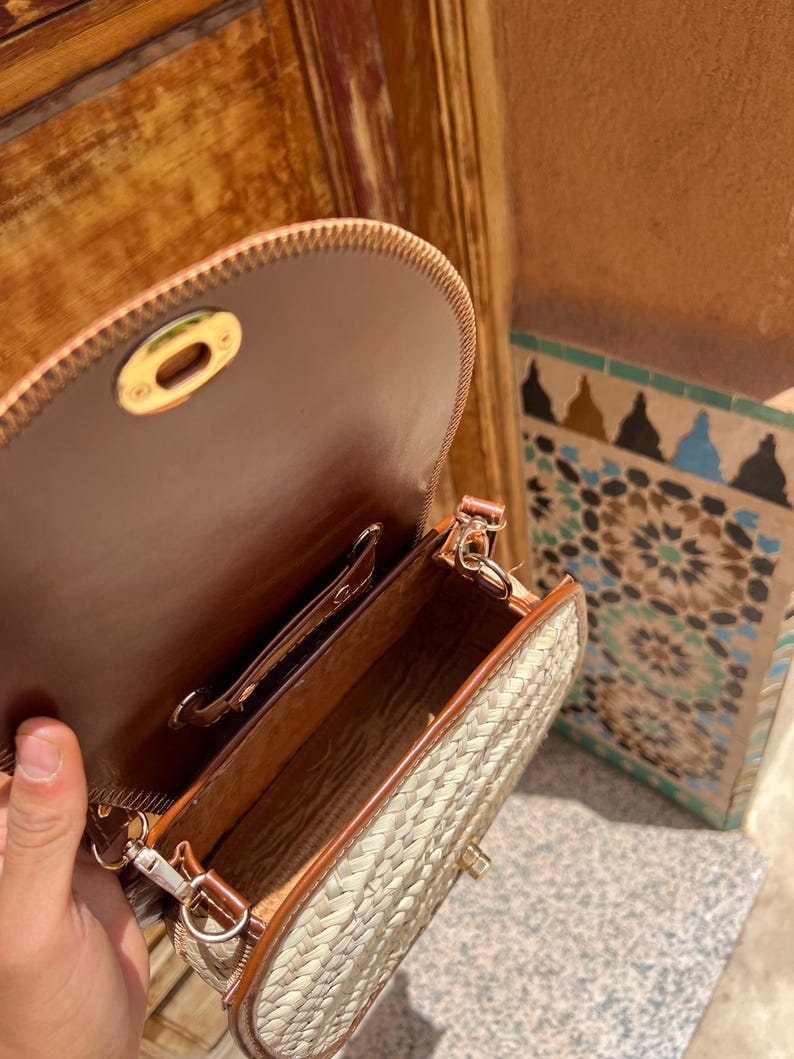 May include: Open view of a woven straw crossbody bag with brown leather trim and a brown leather interior. The bag has a gold-colored clasp and a long, adjustable strap. The bag is partially open, revealing the interior.