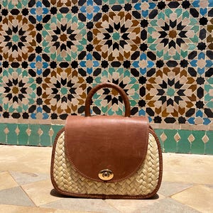 May include: A handcrafted handbag with a woven straw base and a brown leather flap and handle. The bag features a gold-colored clasp and is set against a backdrop of patterned tiles.