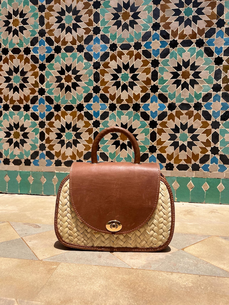 May include: A handcrafted handbag with a woven straw base and a brown leather flap and handle. The bag features a gold-colored clasp and is set against a backdrop of patterned tiles.