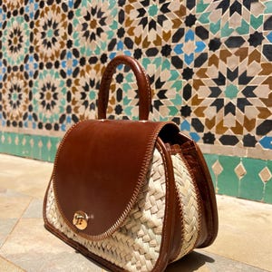 May include: A brown leather and woven straw handbag with a top handle and a gold-colored clasp. The bag has a rounded shape and a woven straw body with brown leather accents.
