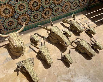 May include: A collection of woven straw animal head sculptures, including a bull, donkey, and other farm animals. The sculptures are in various sizes and are arranged on a light-colored stone surface. The straw is a natural beige color.