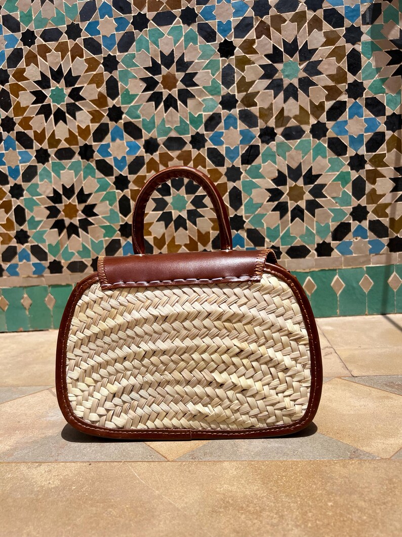 May include: A woven straw handbag with a brown leather top and trim. The bag has a small handle and a rectangular shape. The background features a mosaic tile wall with geometric patterns in shades of blue, brown, and green.