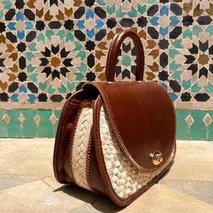 May include: A brown leather handbag with a woven straw design on the sides. The bag has a rounded top with a handle and a gold-colored clasp. The background features a mosaic tile wall.