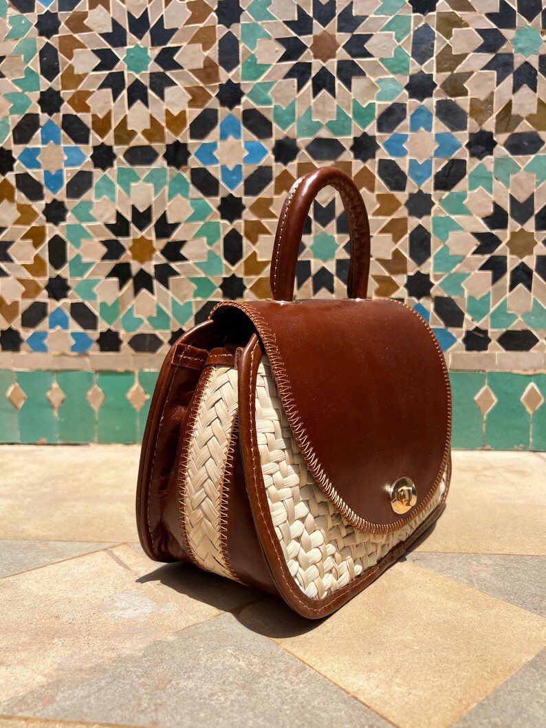 May include: A brown leather handbag with a woven straw design on the sides. The bag has a rounded top with a handle and a gold-colored clasp. The background features a mosaic tile wall.