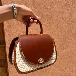 May include: A brown leather and woven straw handbag with a curved top flap and a small handle. The bag has a gold-colored clasp and a woven straw base. The bag is held by a person.