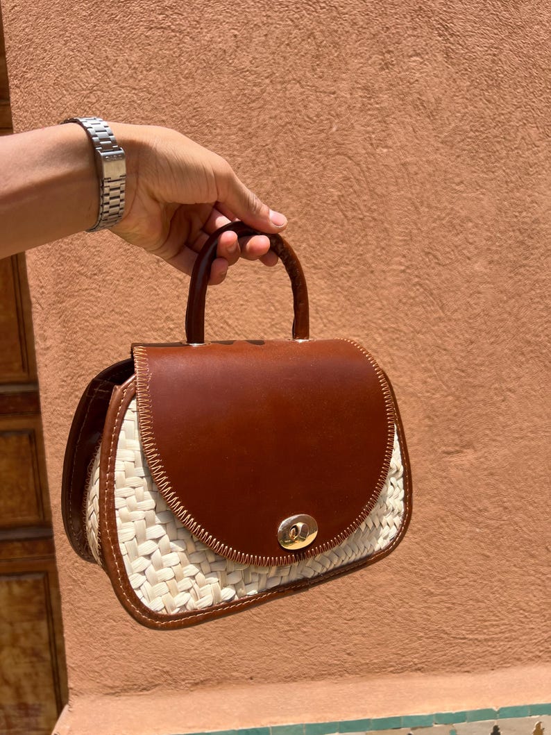 May include: A brown leather and woven straw handbag with a curved top flap and a small handle. The bag has a gold-colored clasp and a woven straw base. The bag is held by a person.