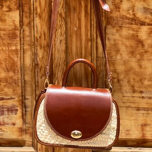 May include: A brown leather handbag with a woven straw base and a top handle. The bag has a flap closure with a gold-colored clasp and a long, adjustable strap. The bag is hanging against a wooden door.