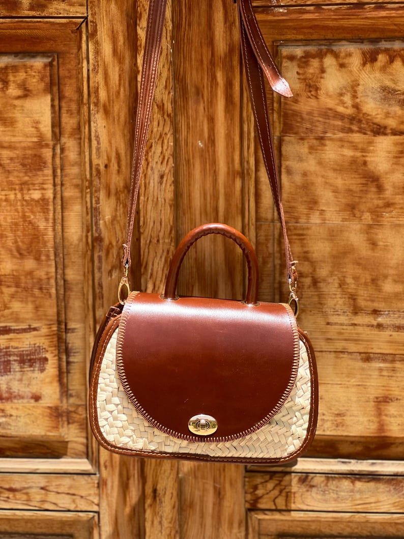 May include: A brown leather handbag with a woven straw base and a top handle. The bag has a flap closure with a gold-colored clasp and a long, adjustable strap. The bag is hanging against a wooden door.