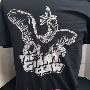 Könnte beinhalten: Schwarzes T-Shirt mit einer weißen Grafik eines vogelartigen Wesens mit ausgebreiteten Flügeln und dem Text "THE GIANT CLAW". Die Website-Adresse "www.AardvarkTees.com" ist unten in Pink aufgedruckt.