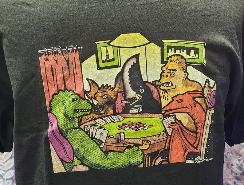 May include: A black t-shirt with a colorful illustration of four monsters playing cards. The monsters are a green dinosaur, a red dragon, a black sea monster, and a brown furry creature. The text "Good As Gold" is printed on the bottom right corner of the shirt.
