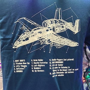 May include: A blue t-shirt with a white outline of an A-10 Warthog aircraft with numbered parts labeled with humorous names. The text reads: 1. BRRT BRRTS, 2. Titanium Man-Tub, 3. Lifty Thingies, 4. GoFasters™, 5. BOOMBS, 6. Drink Holder, 7. Charlie Foxtrotter, 8. Left-Righters, 9. Wobblers, 10. Up-Downers, 11. Meelie-Poppers (not pictured), 12. 10AR BOOOOMBS, 13. Ick's Air Sucker, 14. X cable for listening to 'Highway to the Danger Zone' on repeat, 15. ALEL Sidewinder self defense air-air missiles.