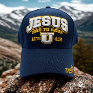 Peut inclure: Casquette de baseball bleu marine avec le texte brodé "JESUS DIED TO SAVE ACTS U 4:12" en jaune et blanc. La casquette est posée sur une roche avec un arrière-plan de montagne.