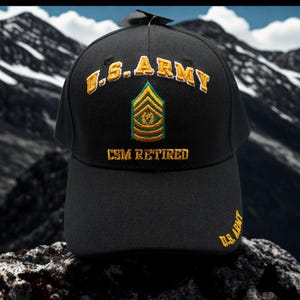 Puede incluir: Gorra de béisbol negra con bordado dorado "U.S. ARMY", una insignia verde y dorada y el texto "CSM RETIRED". La gorra también presenta el texto "U.S. ARMY" bordado en el lateral.