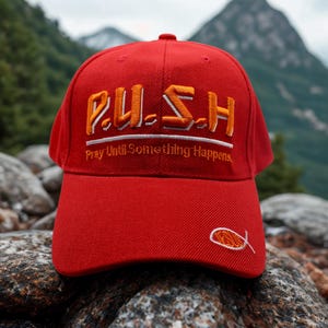 May include: A red baseball cap with the embroidered text "P.U.S.H. Pray Until Something Happens" in orange and white. A small fish symbol is embroidered on the side. The cap is set against a backdrop of mountains and rocks.