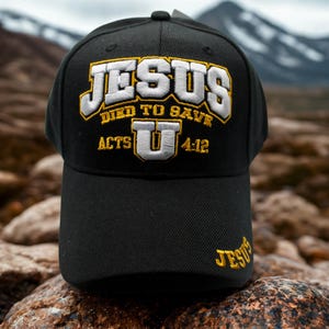 Puede incluir: Gorra de béisbol negra con la palabra "JESUS" bordada en blanco y dorado. El texto "DIED TO SAVE" y "ACTS U 4:12" también están bordados. El fondo es una escena borrosa al aire libre con montañas.