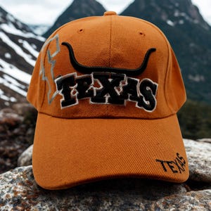 May include: Black trucker hat with a camouflage pattern and a colourful embroidered rooster design. The rooster features yellow, red, and orange details. The hat has a mesh back and a curved brim.