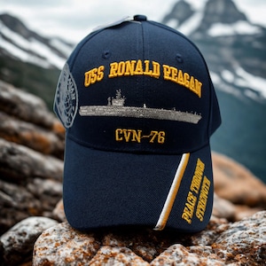 May include: Navy blue baseball cap with gold embroidered text "USS RONALD REAGAN" and a ship illustration. The cap also features "CVN-76" and "PEACE THROUGH STRENGTH" in gold. A patch is visible on the side.