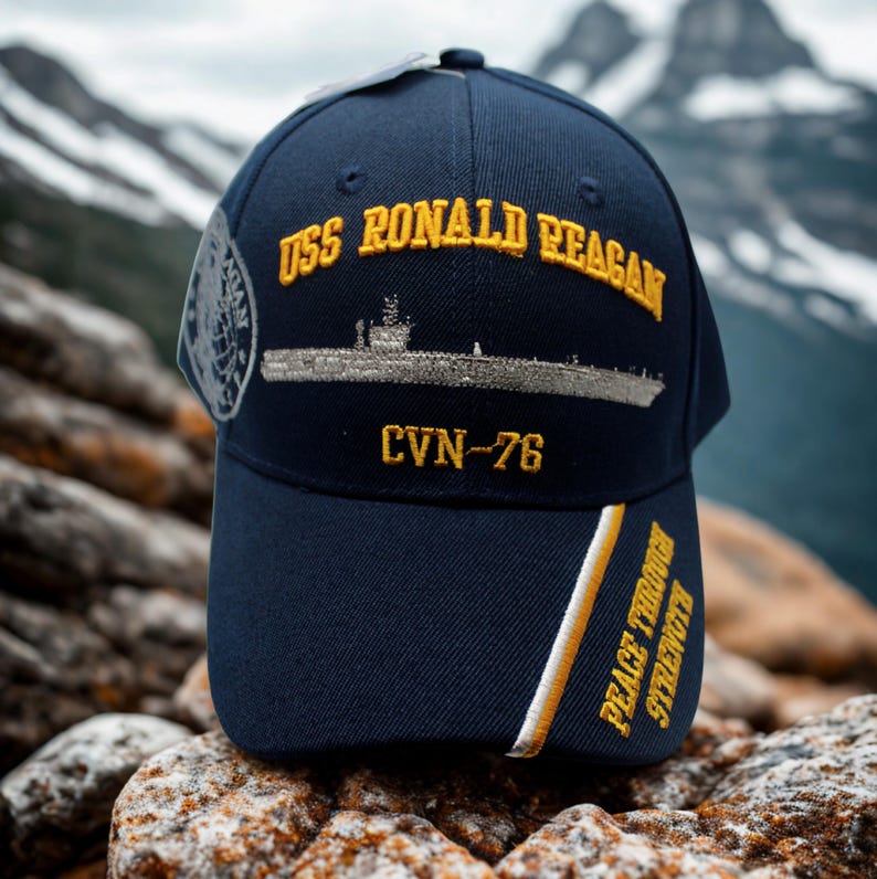 May include: Navy blue baseball cap with gold embroidered text "USS RONALD REAGAN" and a ship illustration. The cap also features "CVN-76" and "PEACE THROUGH STRENGTH" in gold. A patch is visible on the side.