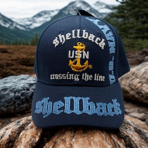 May include: A navy blue baseball cap with embroidered text and a gold anchor emblem. The cap features the words "Shellback", "USN", and "crossing the line" in white and light blue stitching. The cap is set against a natural outdoor backdrop.