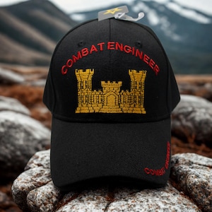 May include: Black baseball cap with "COMBAT ENGINEER" embroidered in red and a gold castle emblem. The cap has a curved brim and a tag attached. The text is also embroidered on the side of the cap.