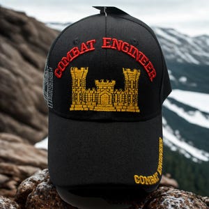 May include: Black baseball cap with "COMBAT ENGINEER" embroidered in red and gold. The cap features a gold castle emblem and additional text on the side. The background shows a mountain range.