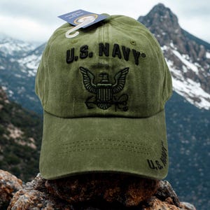 May include: Olive green baseball cap with the text "U.S. NAVY" embroidered in black. The cap features an embroidered emblem of an eagle with a shield. A tag is attached to the cap. The cap is sitting on a rock.