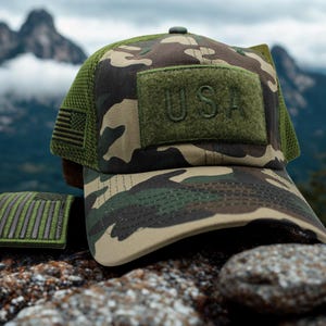Könnte beinhalten: Eine Camouflage-Baseballkappe mit grünem Netzrücken und einem rechteckigen olivgrünen Aufnäher auf der Vorderseite mit dem Text USA. Die Kappe enthält einen kleinen US-Flaggenaufnäher an der Seite und einen passenden Flaggenaufnäher daneben.
