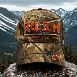 May include: Camouflage baseball cap with the word "PUSH" embroidered in orange, along with the phrase "Pray Until Something Happens." A small fish emblem is on the side. The cap is set against a backdrop of mountains.