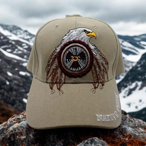 Könnte beinhalten: Eine beigefarbene Baseballkappe mit einer Stickerei eines weißen Adlers mit gelbem Schnabel, der über einem kreisförmigen Schild mit braunen und schwarzen Mustern platziert ist. Der Text NATIVE PRIDE ist in Weiß auf der rechten Seite des Schirms gestickt.