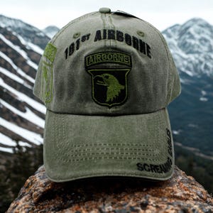Puede incluir: Gorra de béisbol verde oliva con la inscripción "101st Airborne" y un parche con un emblema de águila. La gorra tiene una visera curva y las palabras "Screaming Eagles" están bordadas en el lateral. Montañas en el fondo.