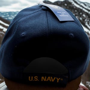 May include: A navy blue baseball cap with a curved brim. The cap features the text "U.S. NAVY" embroidered in gold on the back. A tag is attached to the cap with official branding.